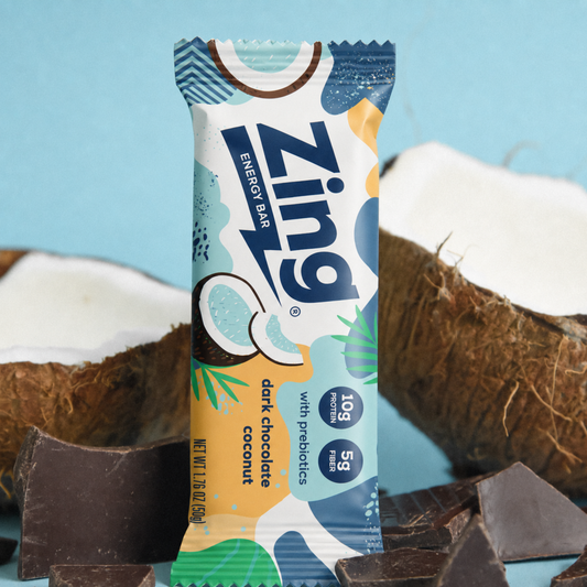 Zing energy bar with dark chocolate and coconut flavor packaging, surrounded by coconuts and chocolate pieces on a light blue background.